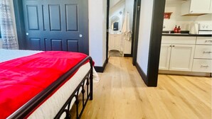 A bedroom with a red bedspread, a wooden floor, a dark door, and a view into a kitchen area with white cabinets. - Little Red Loft | Cozy Guest Suite Apartment (Danville)