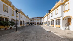 Une cour avec un sol pavé, des bâtiments blancs aux ornements jaunes et des plantes en pot.