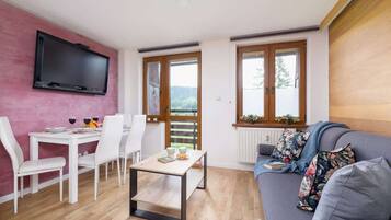 A modern living room with a dining area, a flat-screen TV mounted on a pink wall, a grey sofa with floral cushions, and large windows offering a view of greenery.