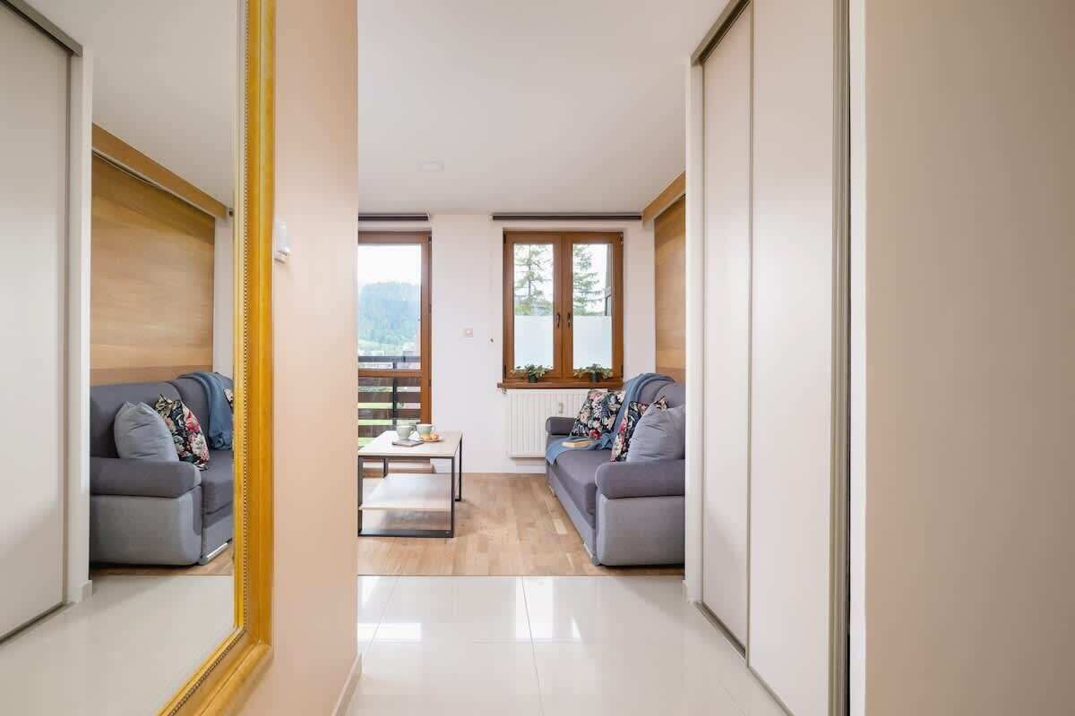 A narrow hallway with two grey sofas and a small table, leading to a window with a view of trees.