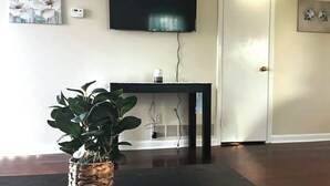 A living room with a large flat-screen TV mounted on the wall, a small table with a decorative item, and a potted plant in the foreground.