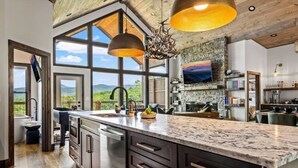 A modern kitchen with a large island, a stone fireplace, and a view of the mountains. - Loaded Luxe w/Pool*Theater*Hot Tub*Fireplace*Game Room*Views! (Sevierville)