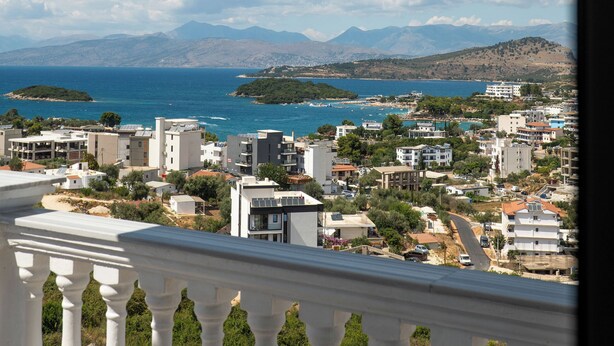 A balcony with white balustrades overlooking a coastal town with buildings and mountains in the background. - Hotel Horizon Ksamil (Ksamil)