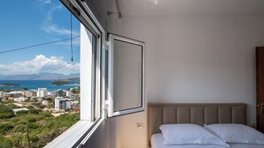 A bedroom with a bed, a window with an open curtain, and a view of a coastal cityscape. - Hotel Horizon Ksamil (Ksamil)