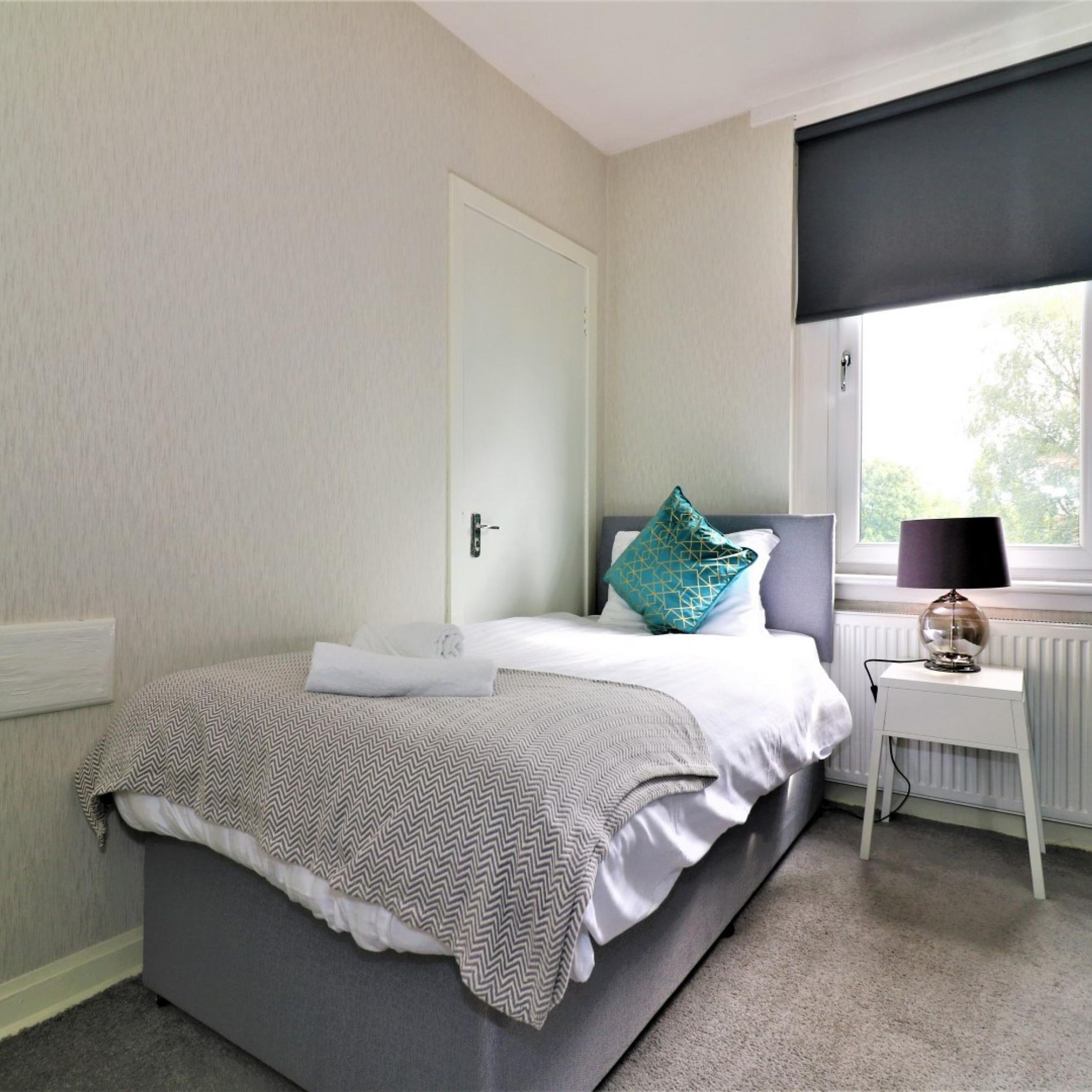 Un dormitorio ordenado con una cama, una mesita de noche con lámpara, una ventana con vistas y una puerta.