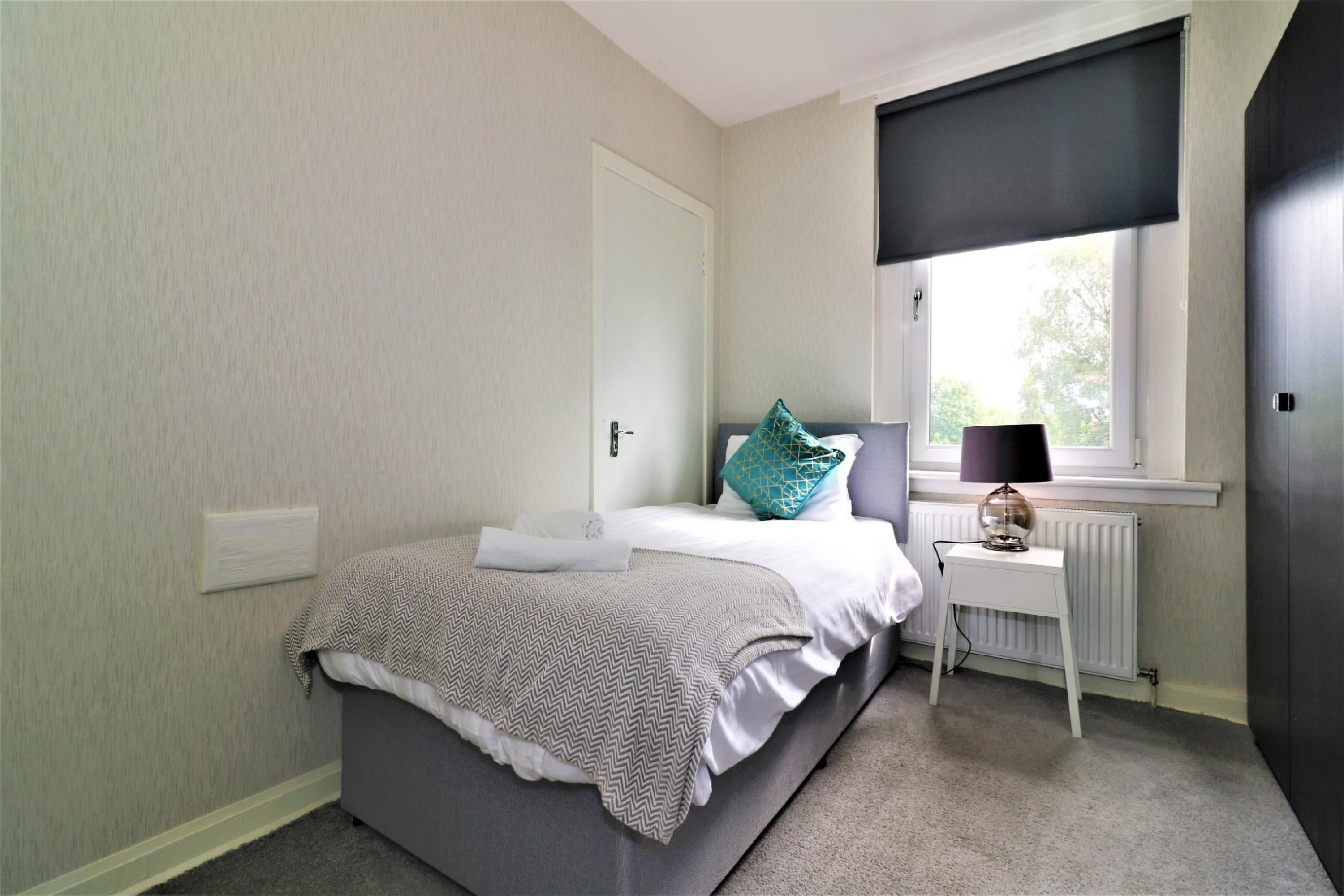 Un dormitorio con una cama, una mesita de noche, una lámpara y una ventana con persiana.