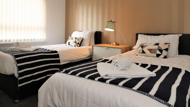 A hotel room with two beds, each with striped bedding, a bedside table with a lamp, and a window with blinds. - Troon House - East Kilbride (Glasgow)