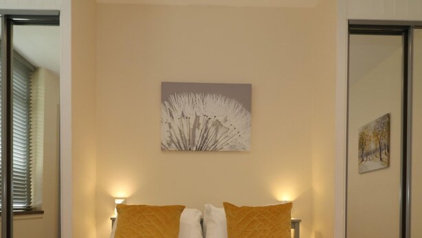 Quarto de hotel com cama, duas almofadas, um quadro na parede e uma janela com persianas.