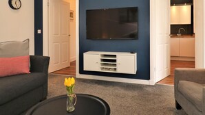 Uma sala de estar com parede azul escuro, uma televisão de tela plana montada acima de um móvel branco, um sofá cinza com almofada vermelha e uma pequena mesa com um vaso de flores.