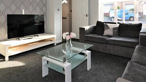 Ett modernt vardagsrum med en platt-TV på ett vitt TV-bord, ett glasbord, en grå soffa med kuddar och en vägg med ett mönster.