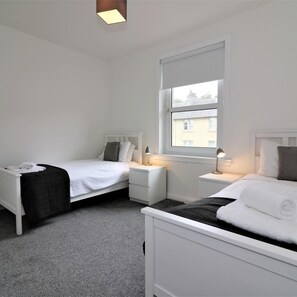 A hotel room with two single beds, a window with a view of buildings, a bedside table with a lamp, and a small bedside table with a phone. - Shawburn House Hamilton (Hamilton)
