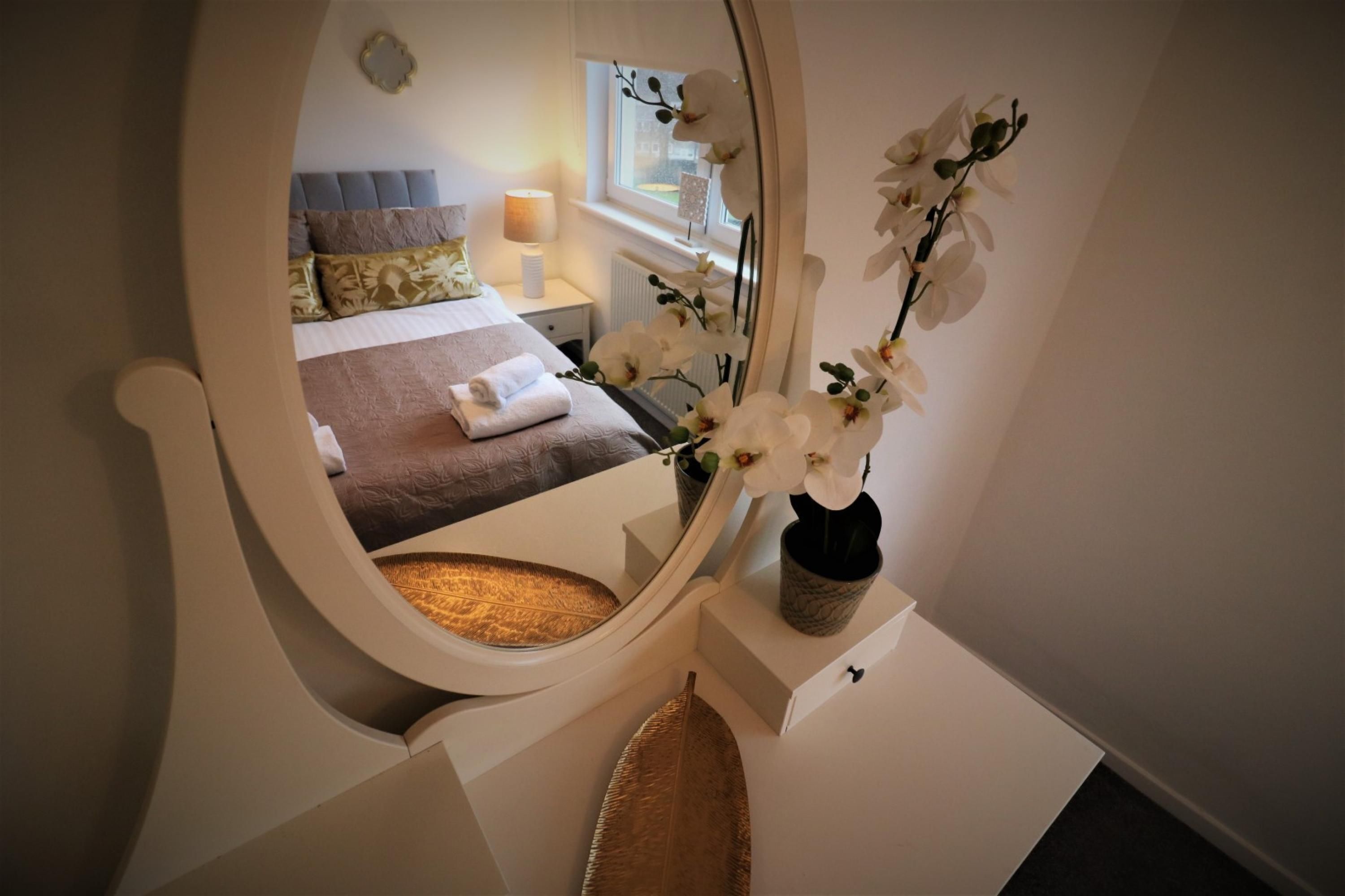 Un dormitorio con cama, una mesita de noche con un florero, un espejo y una ventana.