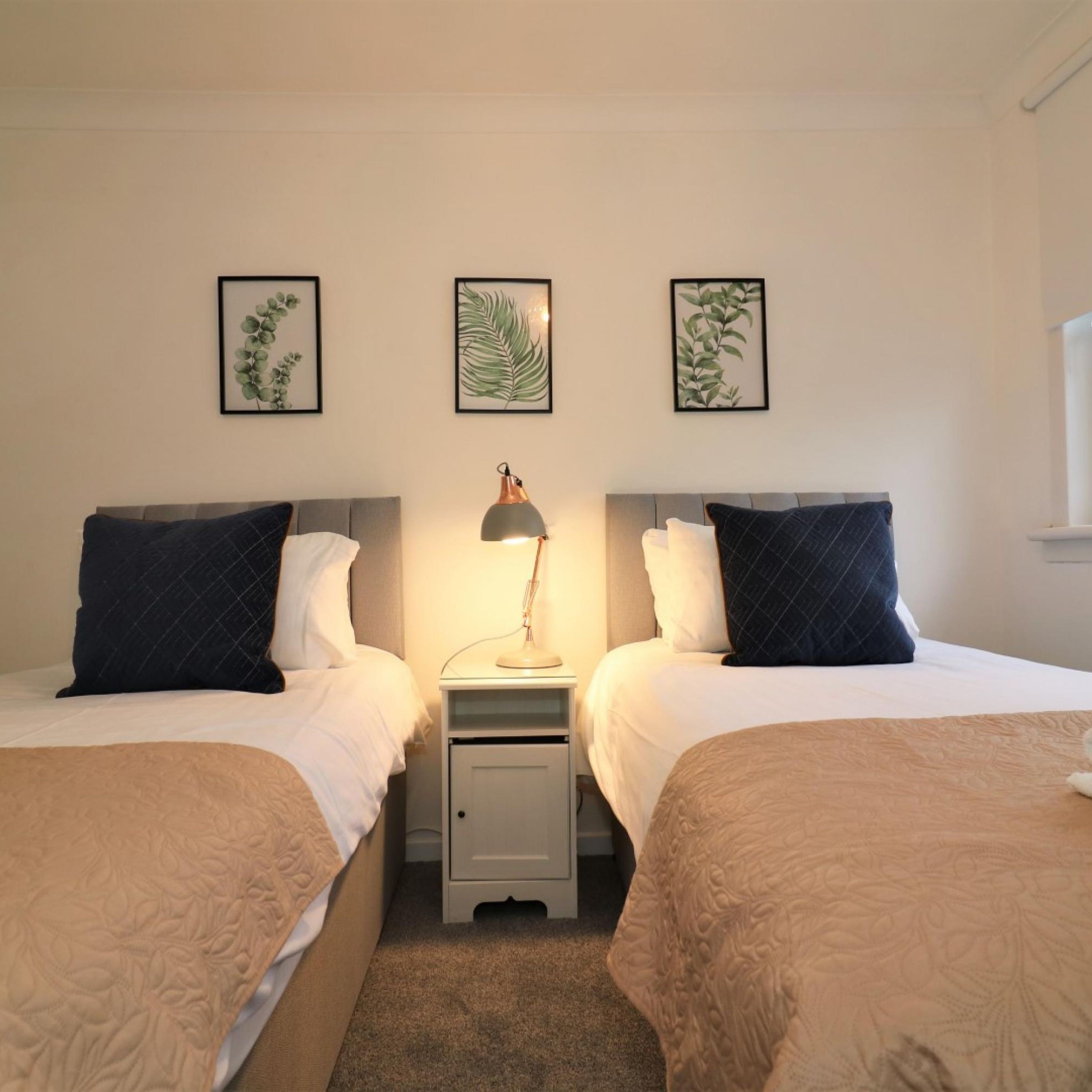 A double room with two beds, a nightstand with a lamp, and framed botanical prints on the wall.