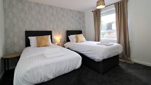 A hotel room with two beds, a window with curtains, a patterned wall, and a nightstand with a lamp.