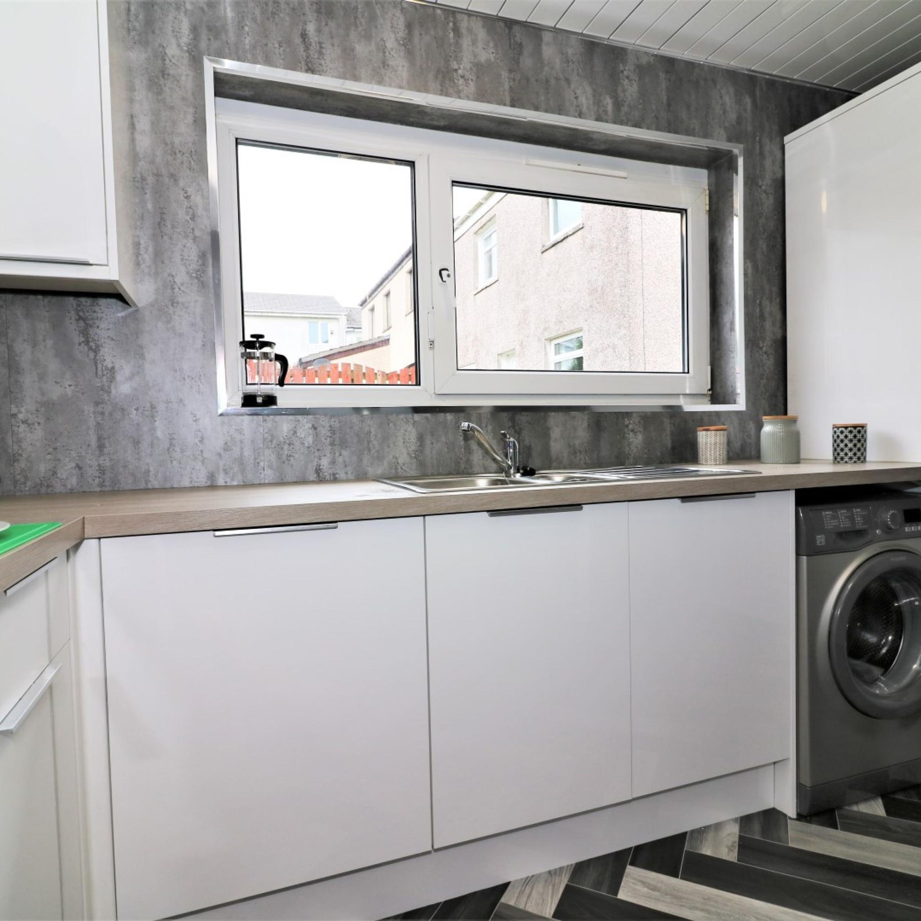 Une cuisine moderne avec des armoires blanches, un lave-linge encastré et une fenêtre donnant sur un quartier résidentiel.