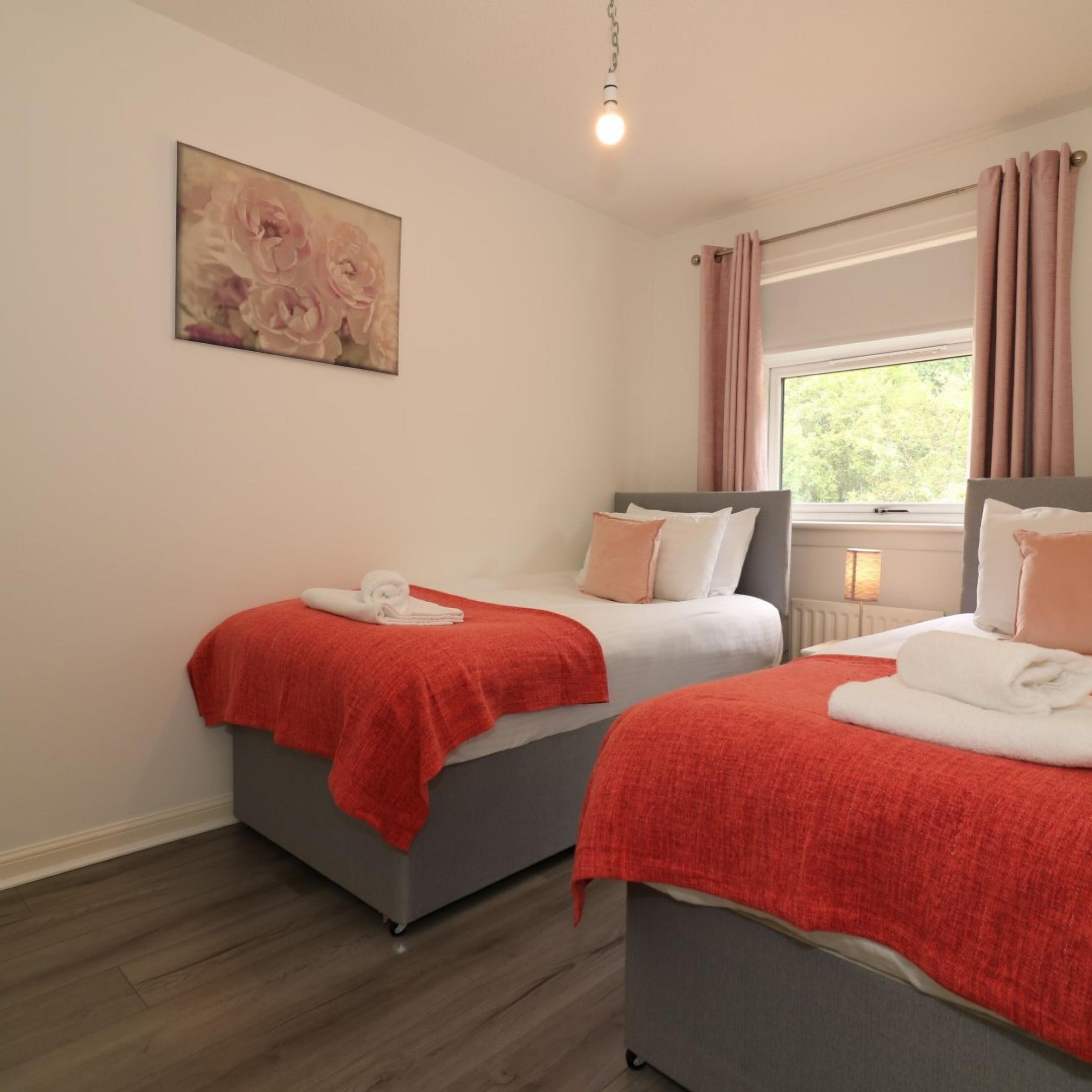 A hotel room with two beds, each with a red blanket and white pillows, a painting on the wall, and a window with curtains.