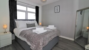 Een slaapkamer met een bed, nachtlampjes, een groot raam met gordijnen, een spiegel en een lijst met een afbeelding aan de muur.