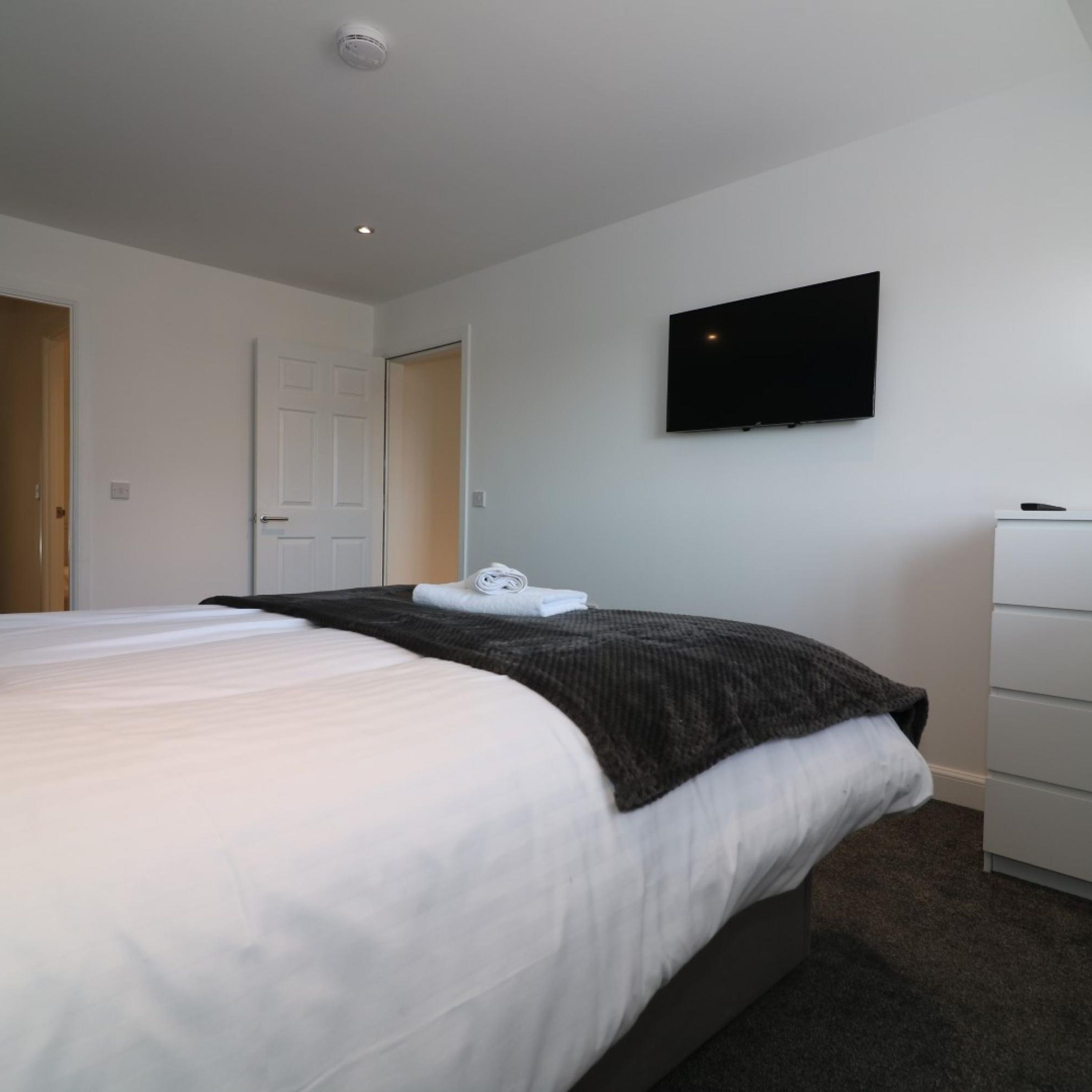 A hotel room with a large bed, a flat-screen TV mounted on the wall, a white dresser, and a door leading to another room.