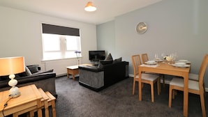 Una sala de estar moderna con un sofá, una mesa de comedor preparada para cuatro personas, un televisor y una ventana con persianas.