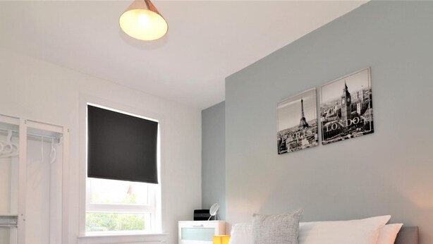 Un dormitorio con cama, ventana, radiador, mesita de noche y una pared con dos fotografías de paisaje urbano en blanco y negro.