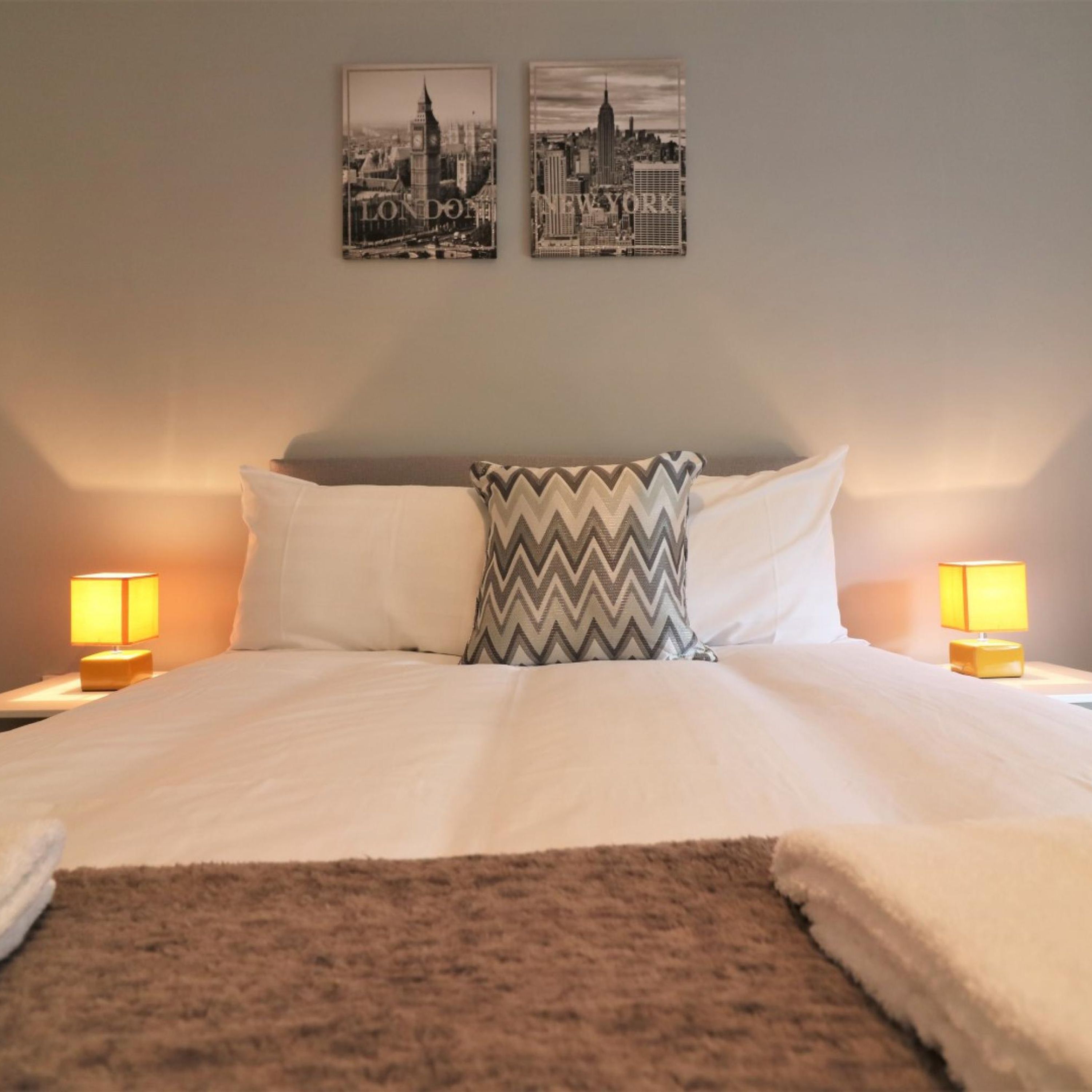 A neatly made bed with white linens, two pillows, a patterned throw, and two bedside lamps. Two framed cityscape photographs are on the wall above the bed.