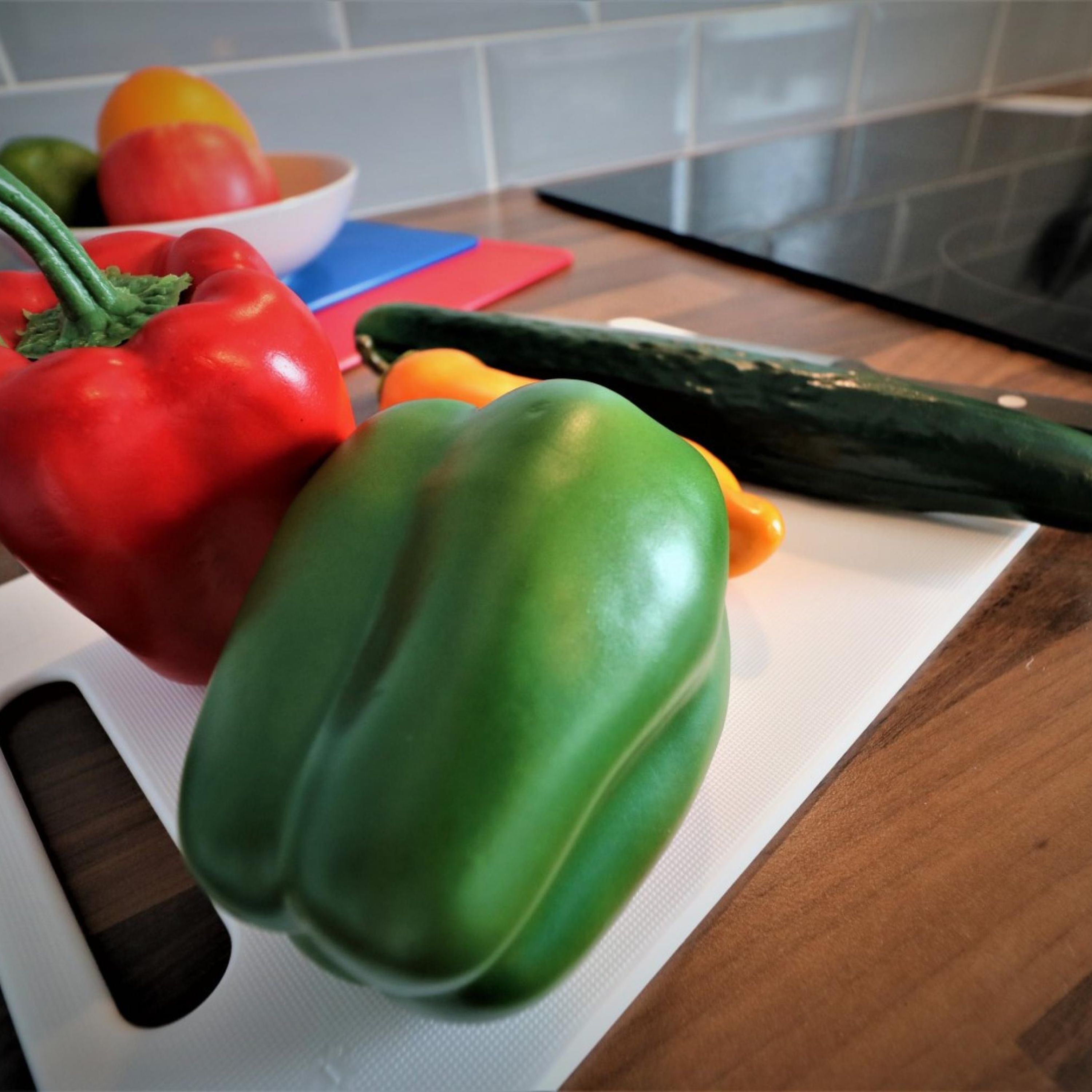Una encimera de cocina con tabla de cortar, un pimiento rojo, un pimiento verde, un pepino y un cuenco con fruta.