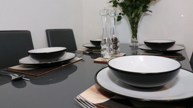Una mesa de comedor dispuesta con platos, fuentes y cubiertos negros, junto a un jarrón de flores y un salero y pimentero.