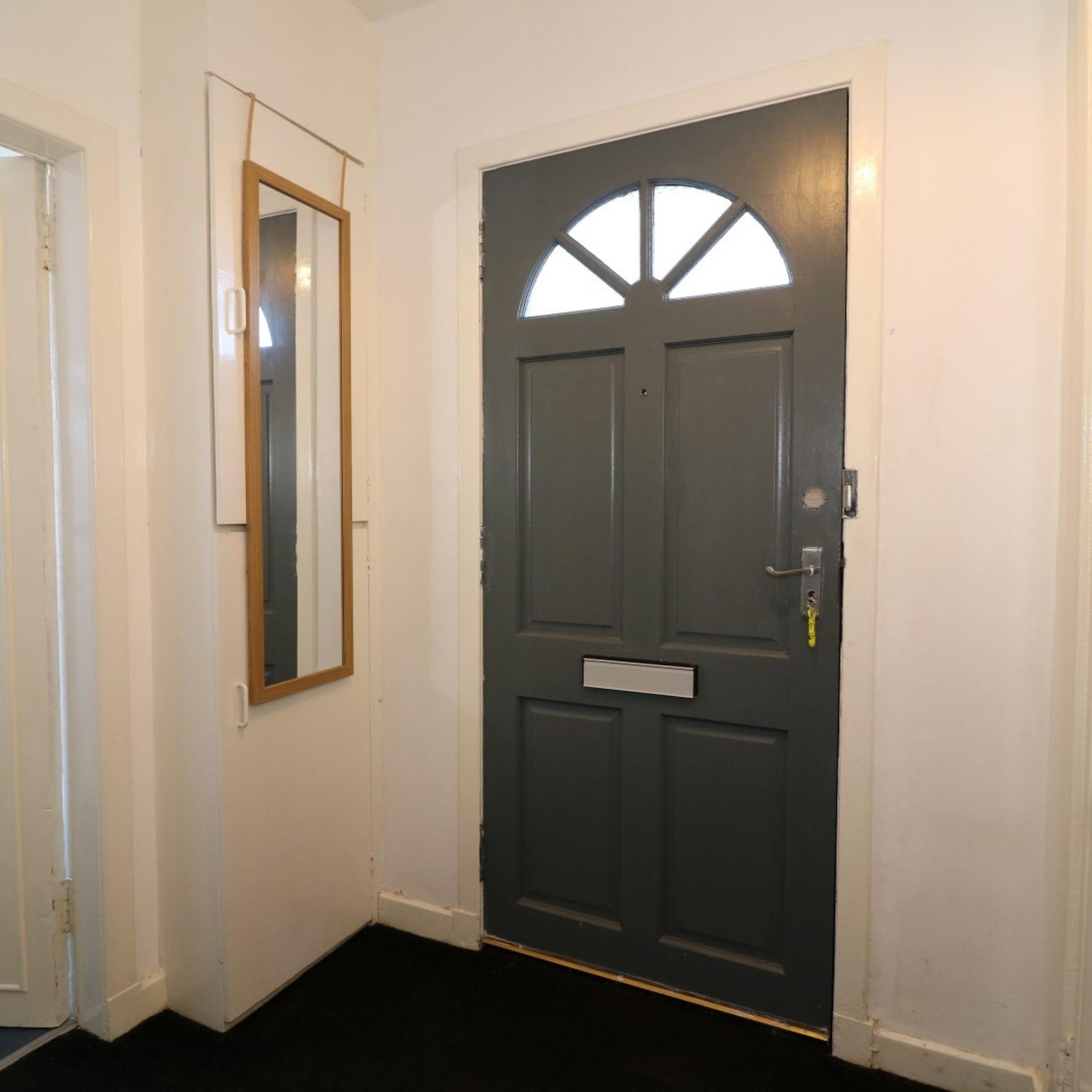 A dark grey door with a small window and a nameplate, white walls, and a mirror on the adjacent wall.