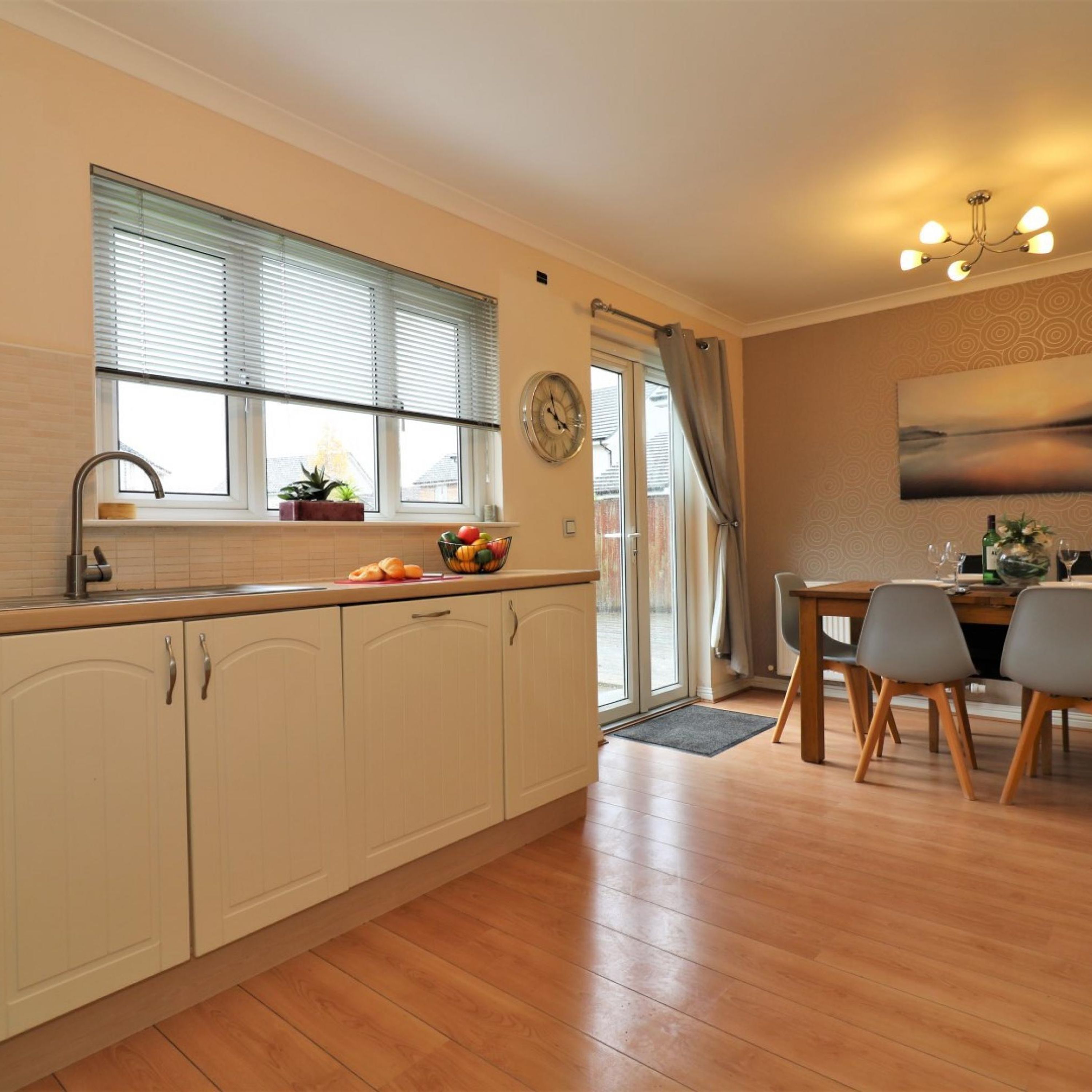 A modern kitchen with a sink, cabinets, and a window with blinds. There is a dining area with a table and chairs.
