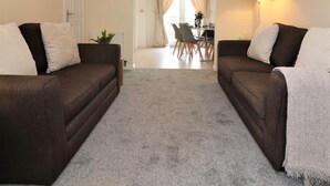 Una sala de estar con dos sofás oscuros, alfombra gris, una araña, un comedor con sillas y una mesa, y una lámpara a la derecha.
