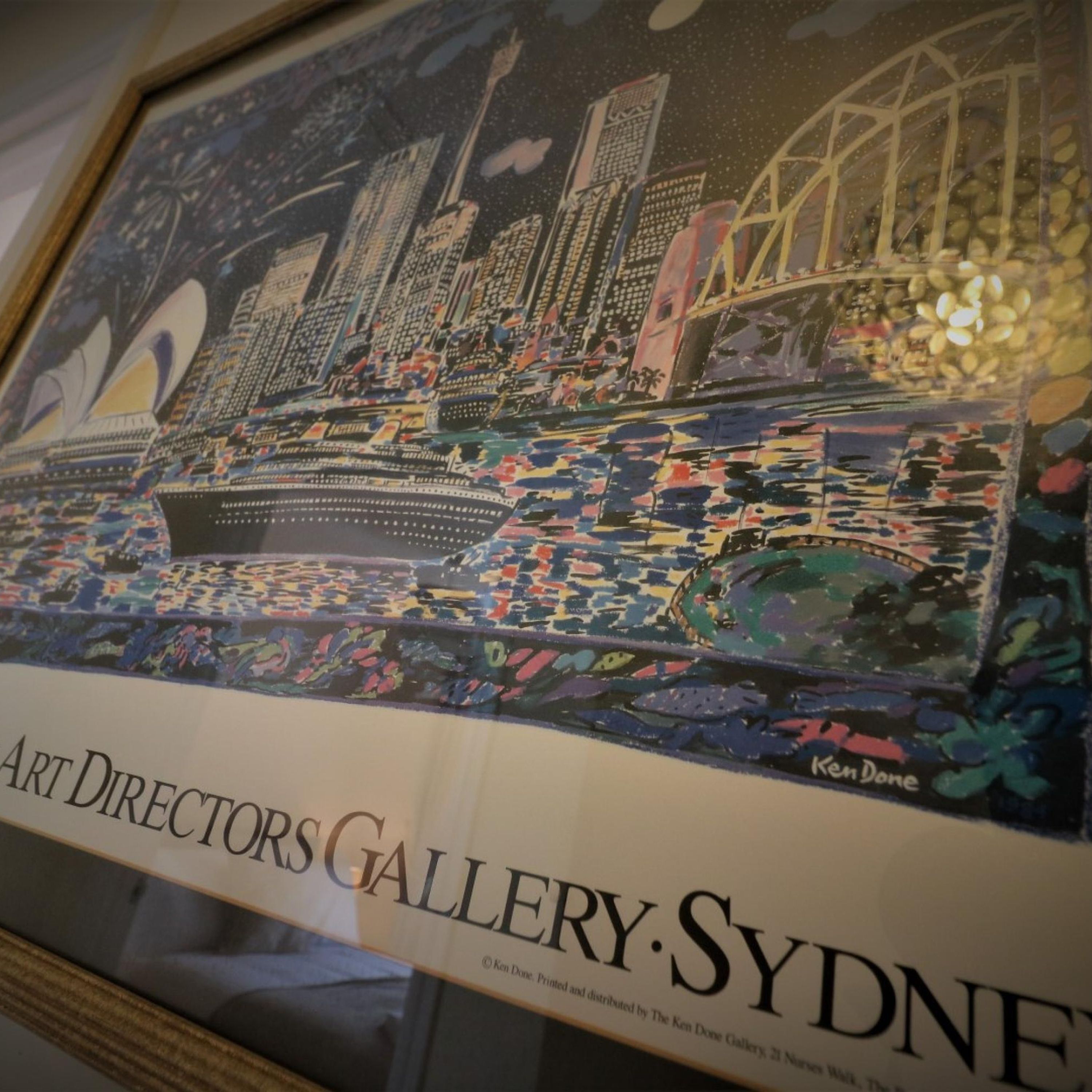 Une œuvre d’art encadrée représentant un paysage urbain avec un navire et un pont, intitulée « Art Directors Gallery Sydney » et réalisée par Ken Done.
