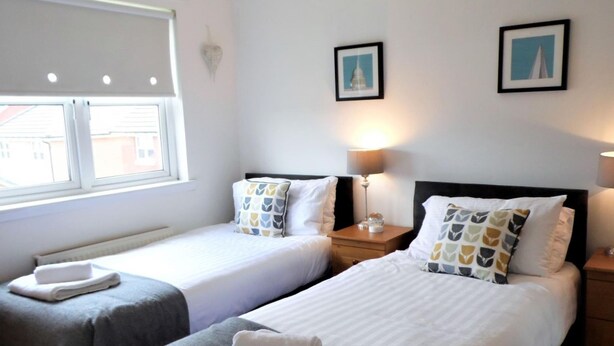 Une chambre d’hôtel avec deux lits simples, un lustre, une fenêtre avec des stores et deux tableaux encadrés accrochés au mur.