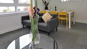 A modern living room with a glass coffee table, a grey sofa, a dining table with yellow chairs, and a large window. - Belhaven House - Hamilton (Hamilton)
