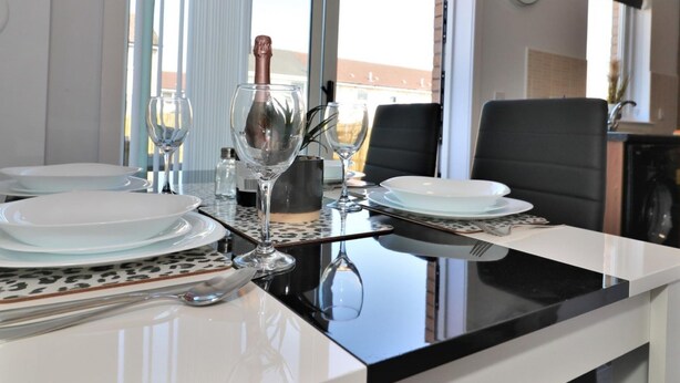 Ein Essbereich mit einem Tisch für zwei Personen, ausgestattet mit weißen Tellern, klaren Weinkellen und einer Champagnerflasche. Der Raum verfügt über große Fenster mit Jalousien, eine Wanduhr und einen Blick auf Gebäude im Freien.