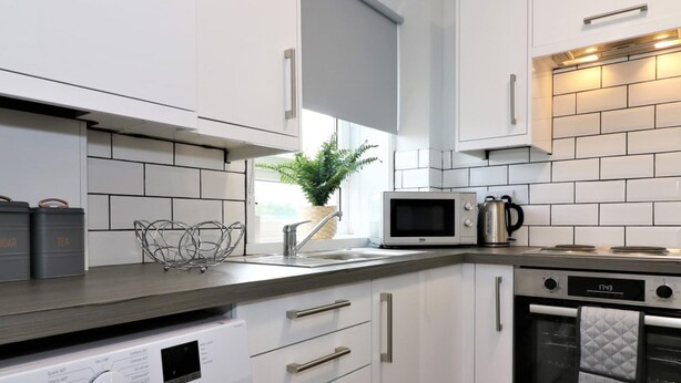 A modern kitchen with white cabinetry, a built-in washing machine, a microwave, and an oven. - Caldercruix House - Airdrie (Airdrie)