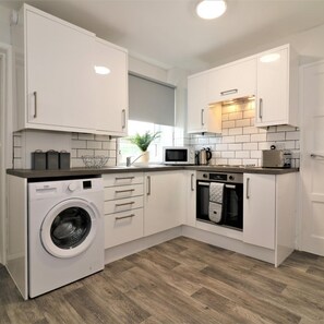 A compact kitchen with white cabinetry, a built-in washing machine, a microwave, and an oven. - Caldercruix House - Airdrie (Airdrie)