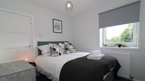 A modern bedroom with a bed, bedside table, and a window with a view of greenery. - Caldercruix House - Airdrie (Airdrie)
