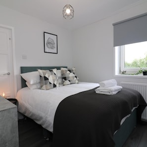 A modern bedroom with a bed, pillows, a bedside table, a framed picture, and a window with a view. - Caldercruix House - Airdrie (Airdrie)