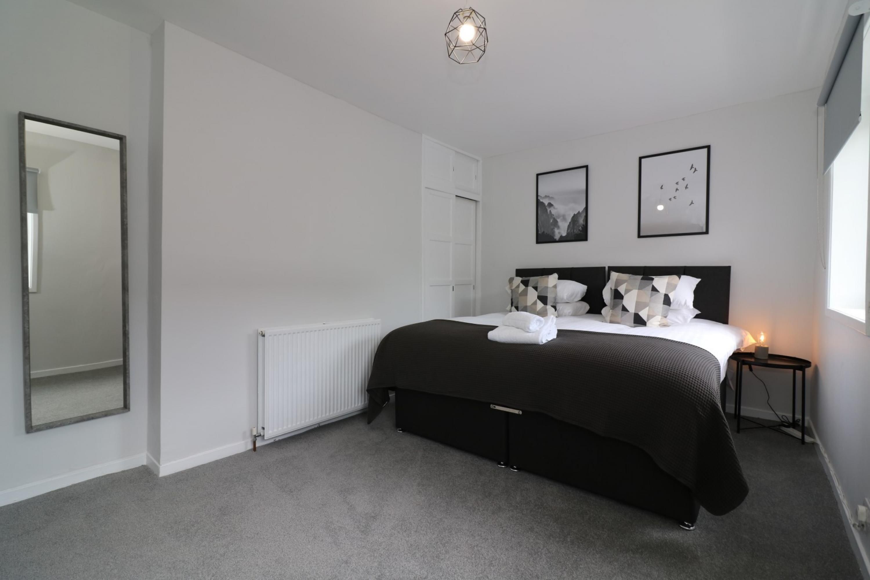 A modern bedroom with a large bed, a bedside table with a lamp, a mirror, and two framed pictures on the wall.