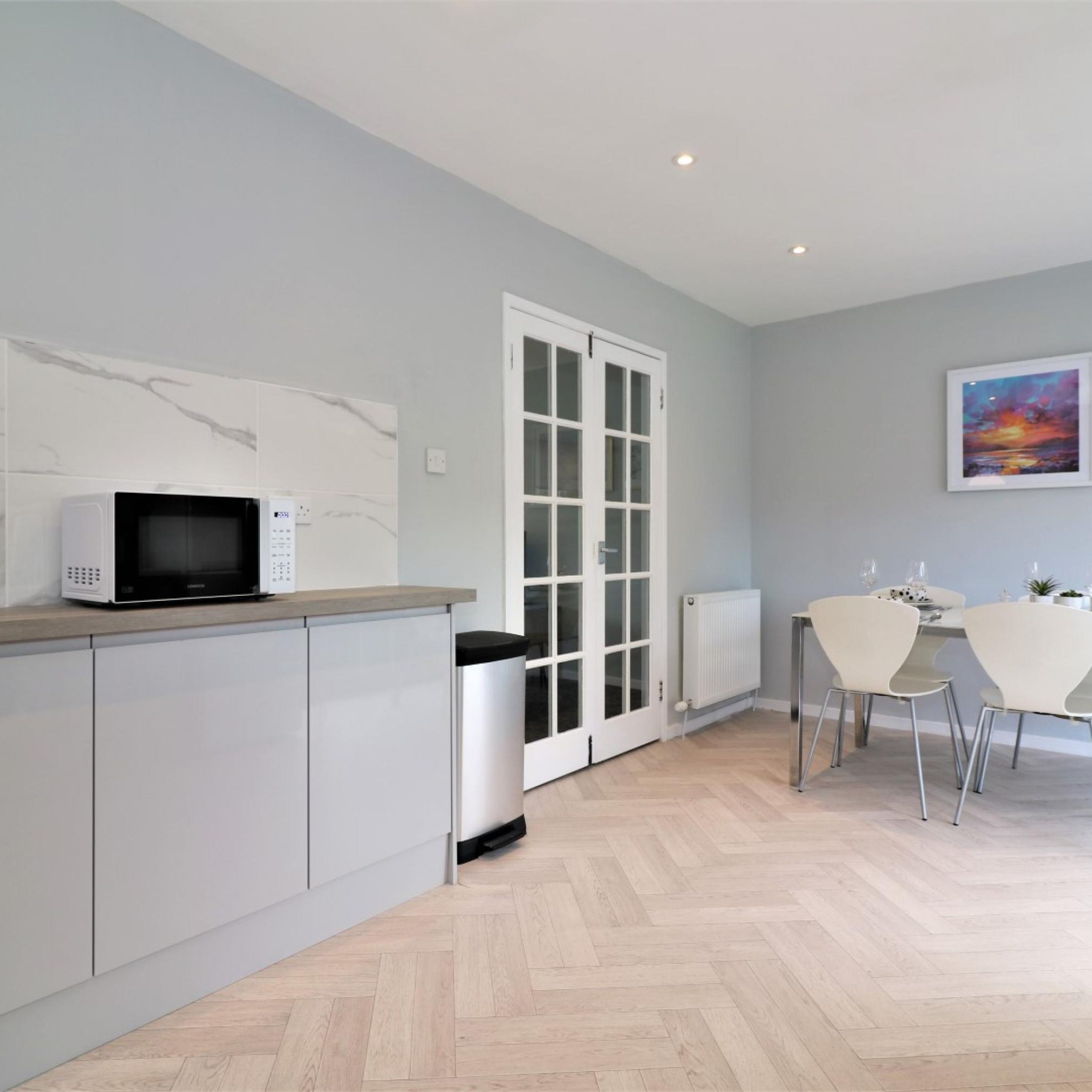 A modern kitchen with a microwave, white cabinets, a white door, and a dining area with two chairs and a table.