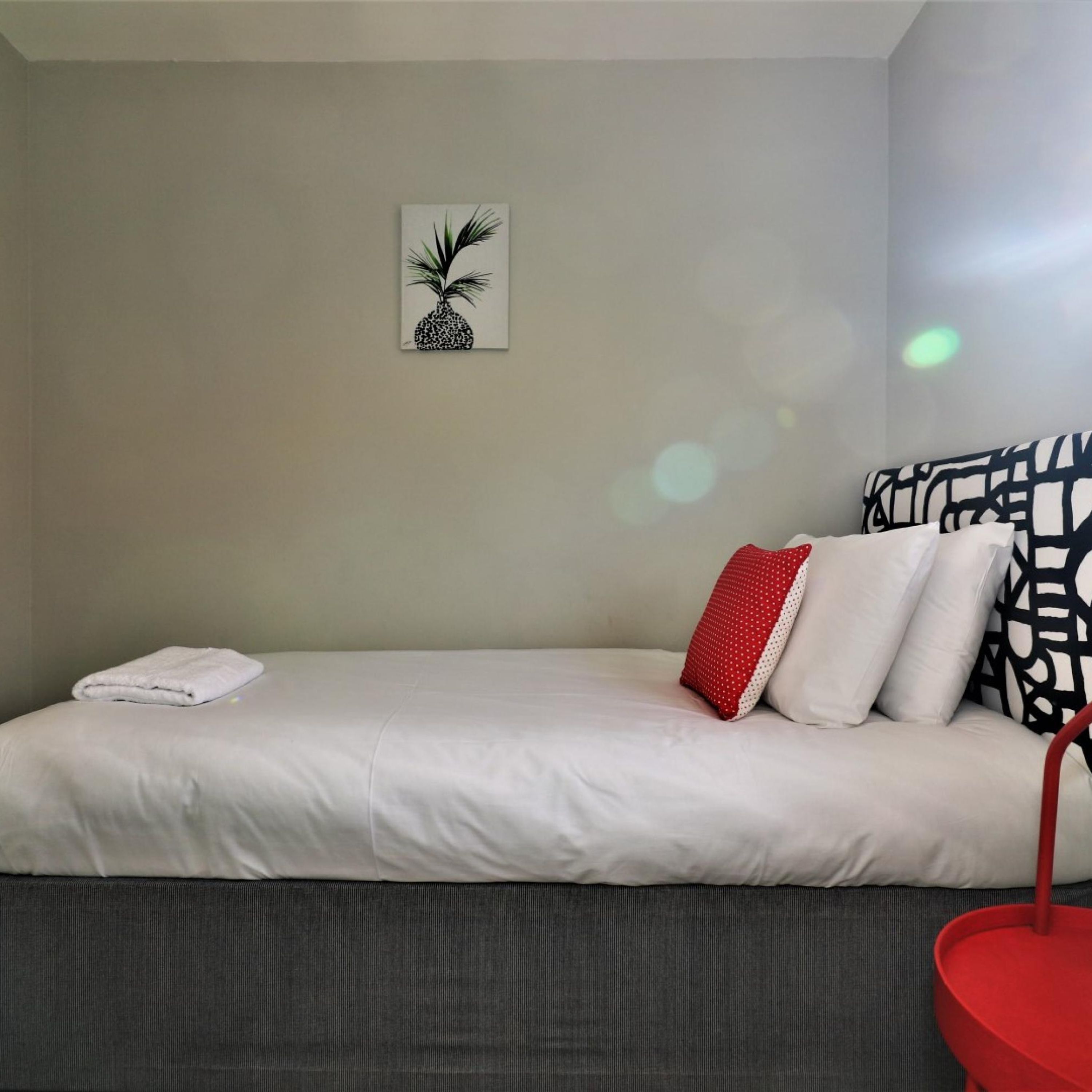 A neatly made bed with white bedding, a red and white pillow, and a pineapple artwork on the wall.