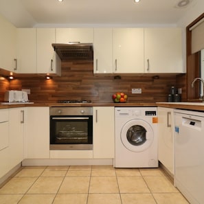 A modern kitchen with white cabinets, a built-in oven, a washing machine, and a wooden backsplash. - Barons Court - Paisley (Paisley)