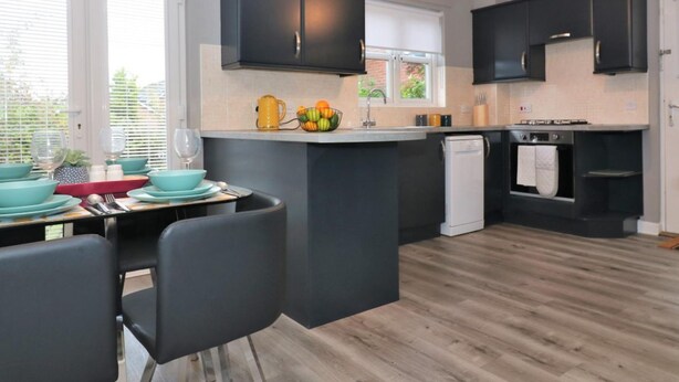 A modern kitchen with dark cabinets, a central island, and a dining area with chairs and a table set for four. - Carnbroe House - Carnbroe (Coatbridge)