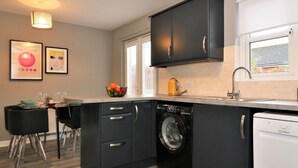 A modern kitchen with black cabinets, a washing machine, and a dishwasher. There is a dining area with a table and chairs, and artwork on the wall. - Carnbroe House - Carnbroe (Coatbridge)