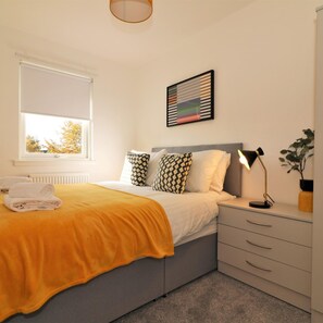 A bedroom with a bed, a bedside table with a lamp, a window with blinds, a radiator, and a framed picture on the wall. - Great Western View - Glasgow (Glasgow)
