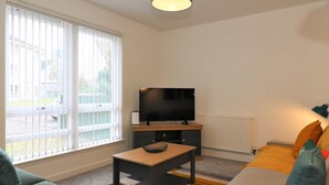 Ein modernes Wohnzimmer mit einem Flachbildfernseher, zwei Sofas, einem Couchtisch und einem großen Fenster mit Jalousien.