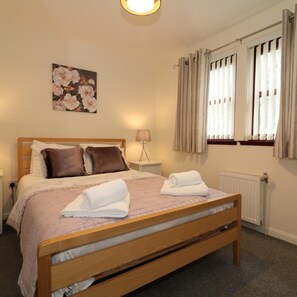 A neatly made bed with a wooden frame, a bedside lamp, and a floral painting on the wall. - Douglas View Blantyre (Glasgow)