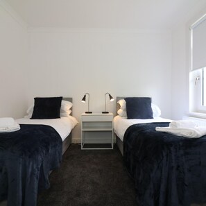 A twin room with two beds, each with a black headboard and white bedding, a small bedside table with a lamp, and a window on the right wall. - New View House Bellshill (Bellshill)