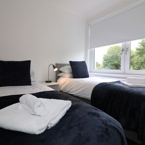 A hotel room with two beds, white walls, a window with a view of trees, and a bedside table with a lamp. - New View House Bellshill (Bellshill)
