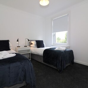 A bedroom with two beds, a nightstand, a lamp, and a window. - New View House Bellshill (Bellshill)